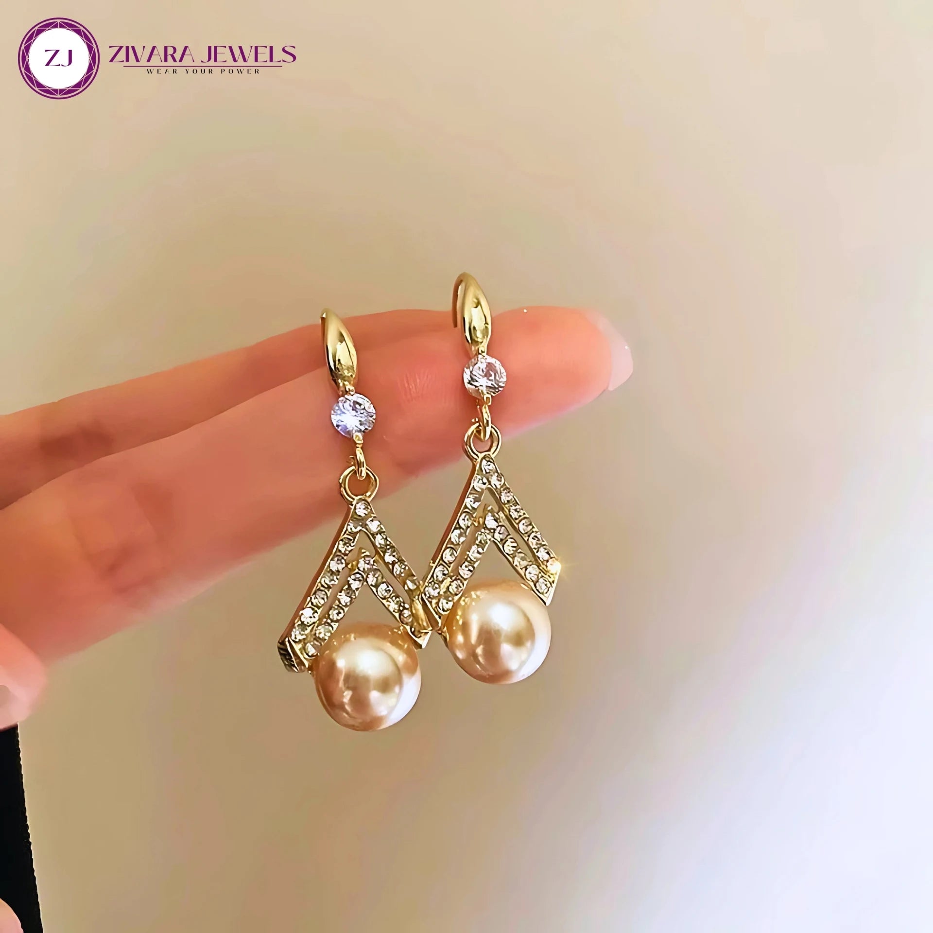 Gold earrings with pearls held by a hand against a blurred background, featuring Zivara Jewels logo.